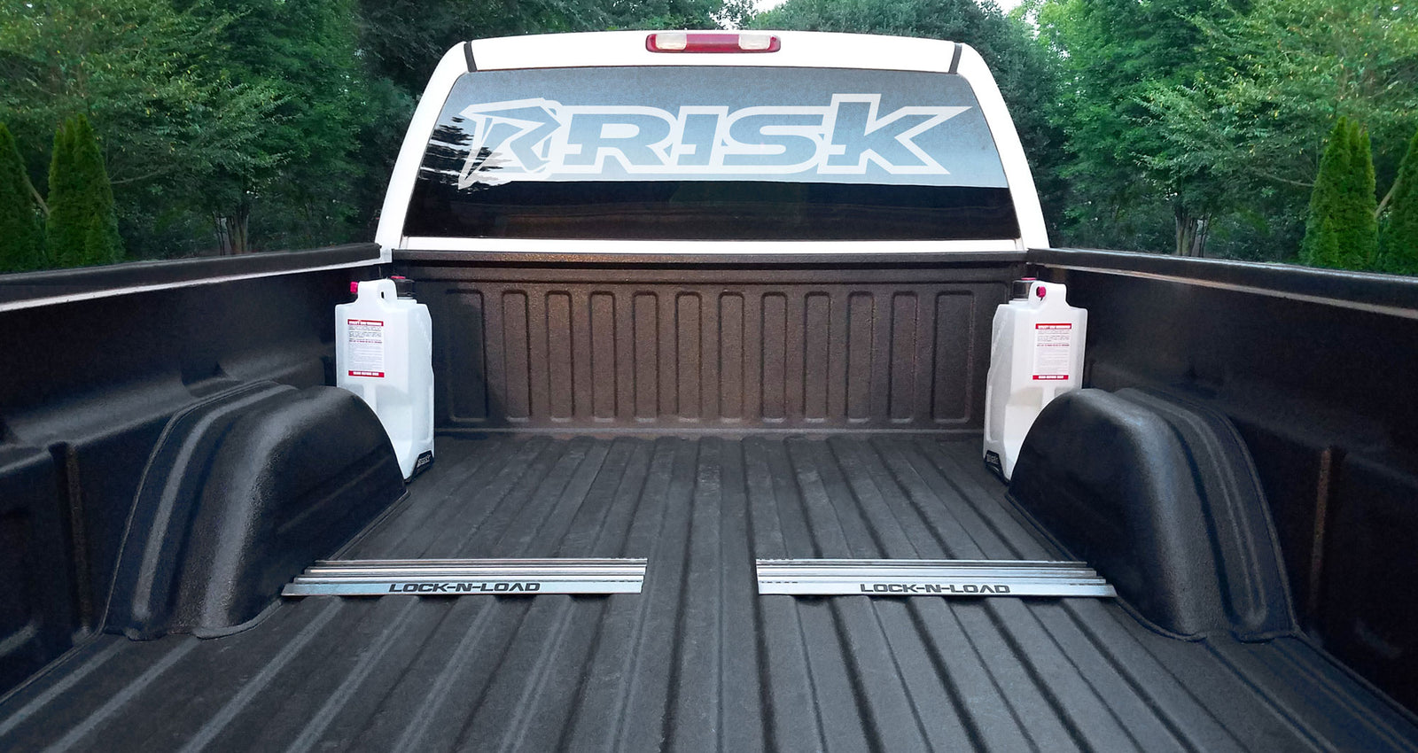 Black spray lined truck bed featuring two Lock-n-load mounting plates installed in between the wheel wells. Also features two ez jugs installed. White truck cab with huge Risk Racing sticker in back window.