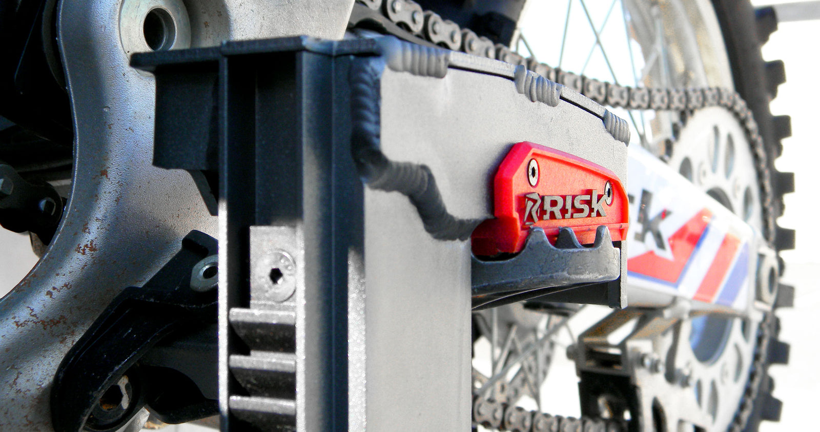 Super close up of the jaw of a lock-n-load pro clamping down on a dirt bike's foot peg. Swing arm and rear tire fading out into the white studio background.