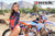 February Moto Model Alliyah wearing a Black & Red Risk Racing MX Jersey and bikini bottoms standing side profile in front of a KTM dirt bike #277 at a MX track with mountains in background.