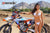 Medium shot of February Moto Model Alliyah wearing a bikini standing in front of a KTM dirt bike #277 sitting on a Risk Racing ATS MX stand at a MX track with mountains in the background. Her right arm draped across the handle bars.