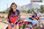 Medium shot of February Moto Model Alliyah wearing a Red & Black Risk Racing MX Jersey and blue bikini bottoms standing in front of a KTM dirt bike #277 at a MX track with mountains in background. Both hands slightly tugging the jersey down.