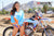 Medium shot of February Moto Model Alliyah wearing a Blue & Orange Risk Racing MX Jersey and blue bikini bottoms standing in front of a KTM dirt bike #277 at a MX track with mountains in background.