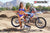 Wide shot of February Moto Model Alliyah in a blue bikini standing in front of a KTM dirt bike #277 sitting on a Risk Racing ATS MX stand at a MX track with mountains in background. Her left hand on the throttle and right on back fender.