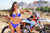 Close up of February Moto Model Alliyah wearing a big smile and a small blue bikini standing in front of a KTM dirt bike sitting on a Risk Racing ATS MX stand at a MX track with mountains in background. Her left arm draped over the throttle.