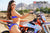 Risk Racing's February Moto Model Alliyah side pose showing off the back of her blue bikini standing in front of a KTM dirt bike at a MX track with mountains in background. Left hand resting on the throttle.