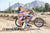 Wide shot of February Moto Model Alliyah wearing a blue bikini standing in front of a KTM dirt bike number plate 277 sitting on a Risk Racing ATS MX stand with her hand resting on a handle bar grip. At a MX track with mountains in background.