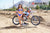 Wide shot of February Moto Model Alliyah wearing a blue bikini standing in front of a KTM dirt bike sitting on a Risk Racing ATS MX stand with one hand resting on a handle bar grip. Other hand on her thigh. At a MX track with mountains in background.