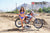 Wide shot of February Moto Model Alliyah wearing a blue bikini standing in front of a KTM dirt bike sitting on a Risk Racing ATS MX stand at a MX track with mountains in background. Left knee up cover right. Left arm across torso. Right hand touching hair