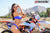 Close up of February Moto Model Alliyah side profile wearing a blue bikini standing in front of a KTM dirt bike #277 at a MX track with mountains in background. Left hand and right elbow on the throttle. Right hand touching her temple