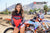Close up of February Moto Model Alliyah tugging her Black & Red Risk Racing MX Jersey down almost covering her blue bikini bottoms standing in front of a KTM dirt bike #277 at a MX track with mountains in background.