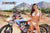 February Moto Model Alliyah in a bikini standing in front of a KTM dirt bike #277 sitting on a Risk Racing ATS MX stand at a MX track with mountains in background. Her right hand on the bikes left handle bar grip