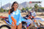 Close up of February Moto Model Alliyah tugging her Blue & Orange Risk Racing MX Jersey down almost covering her blue bikini bottoms standing in front of a KTM dirt bike #277 at a MX track with mountains in background.