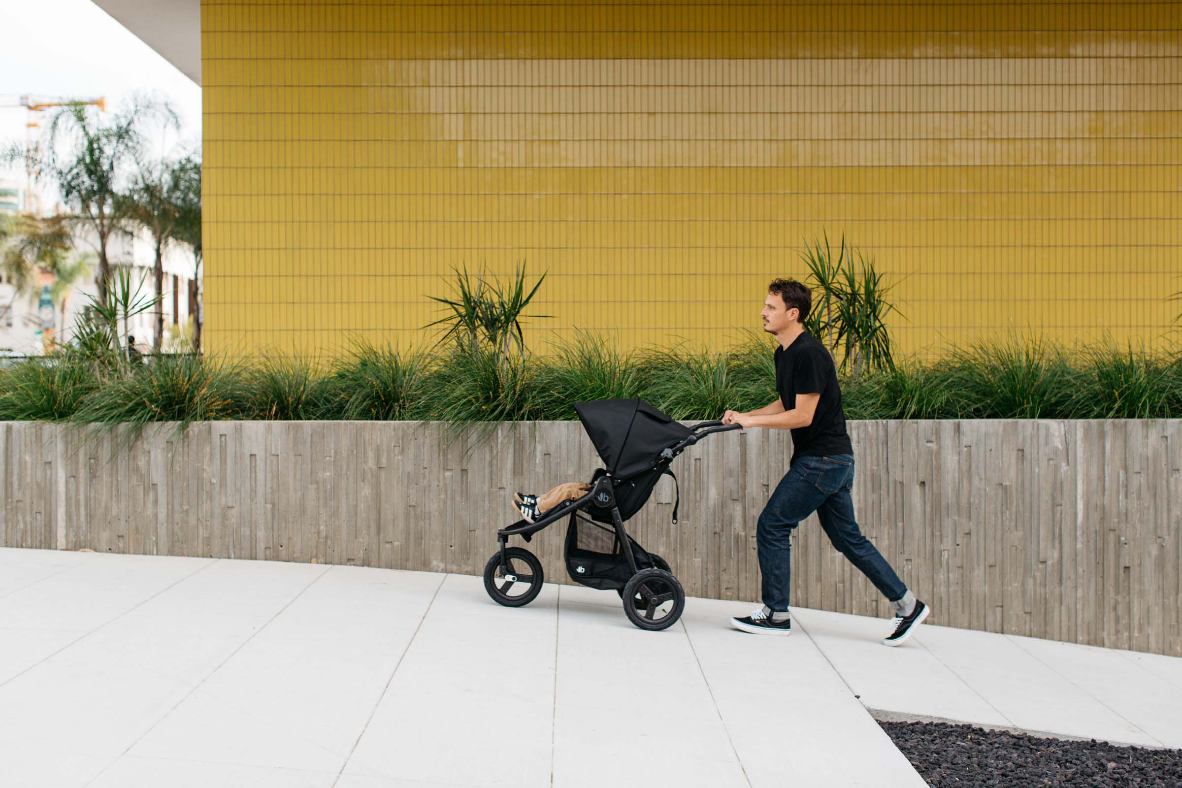 Picture of father pushing black Indie stroller uphill with toddler toes peeking out against a gold wall background in city.
