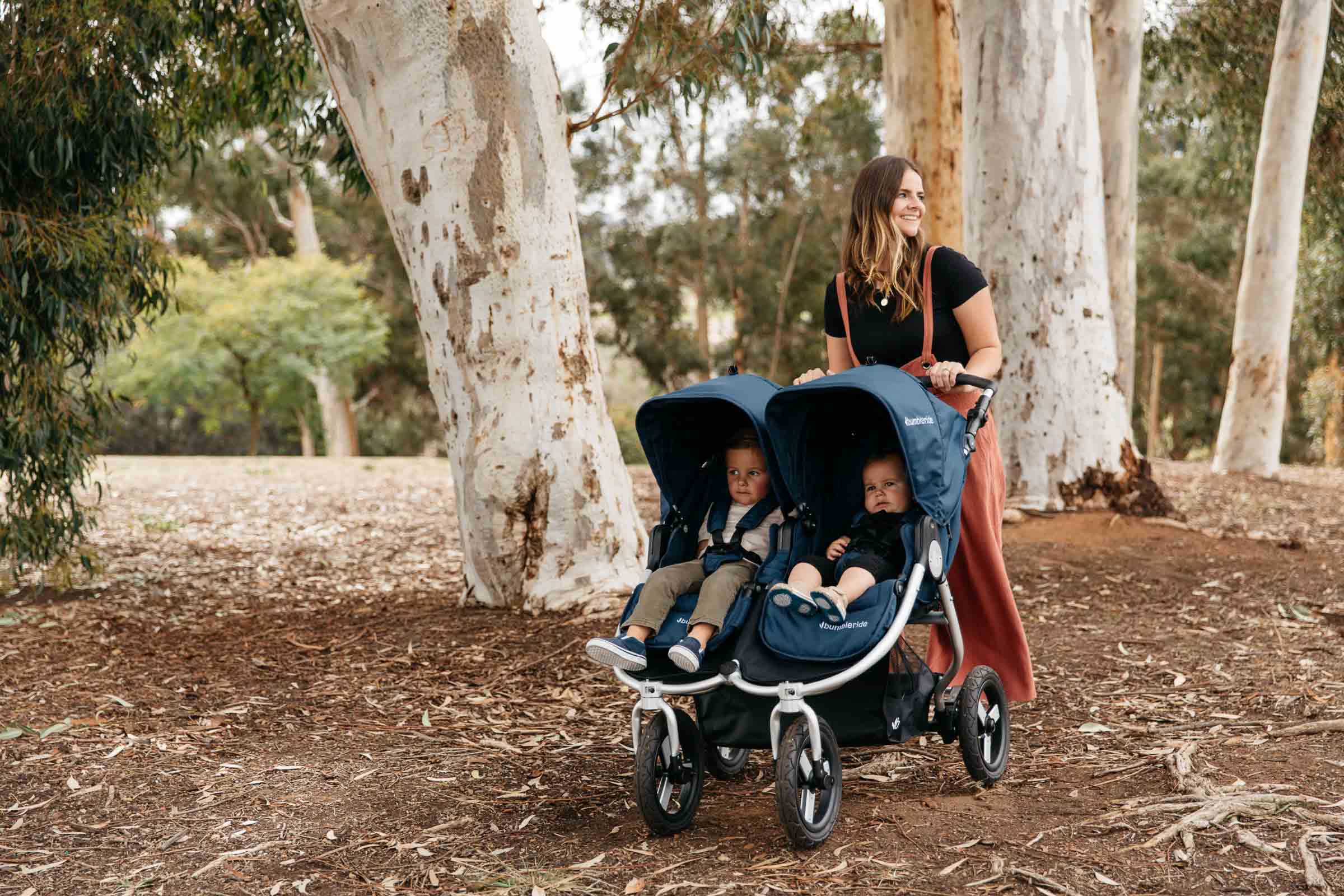Picture of mother smiling over shoulder while pushing double stroller over large mulch with two children riding inside looking into the distance.