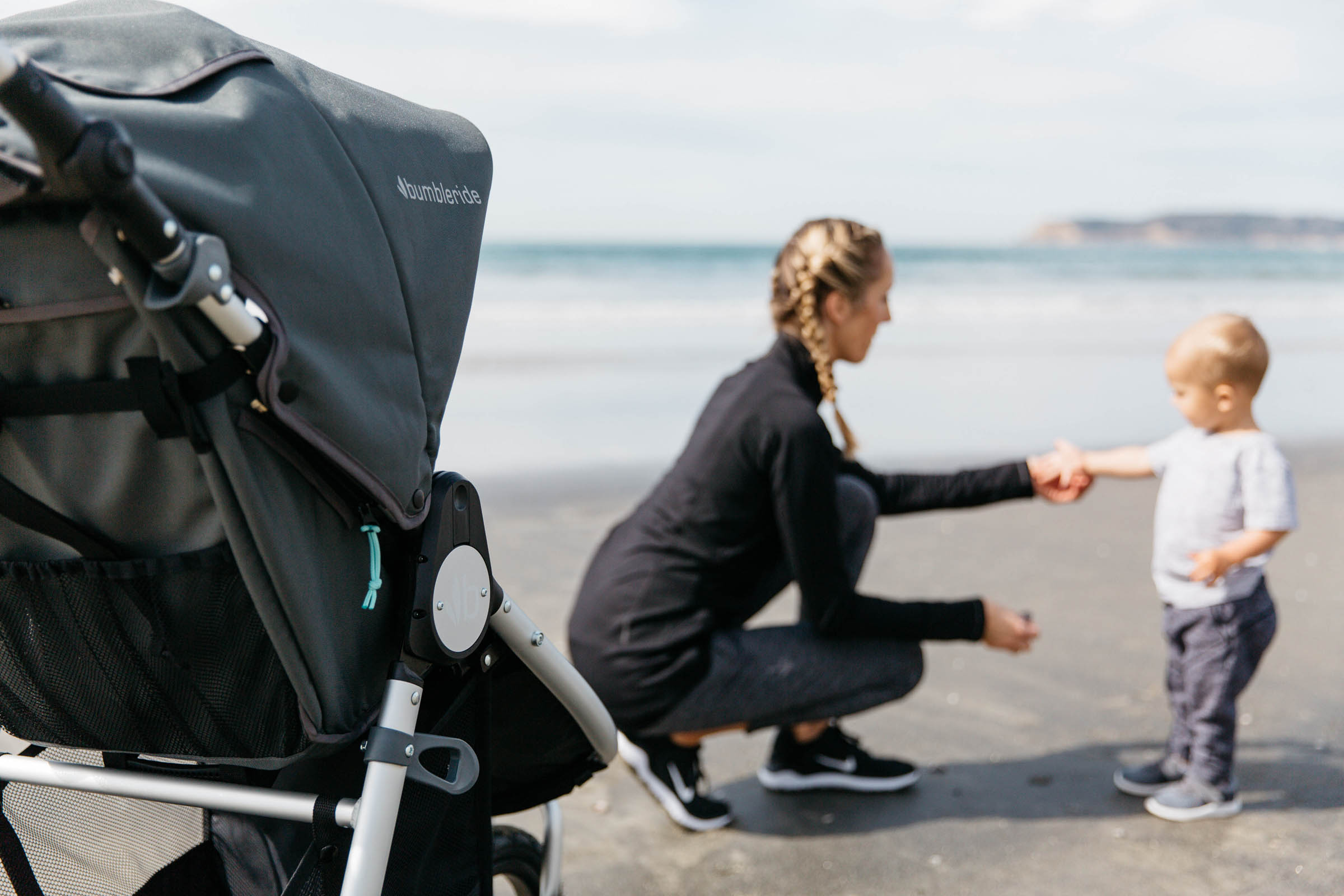 Picture of mother kneeling next to toddler with hand out showing them a shell or similar with Speed stroller in foreground and ocean in background.