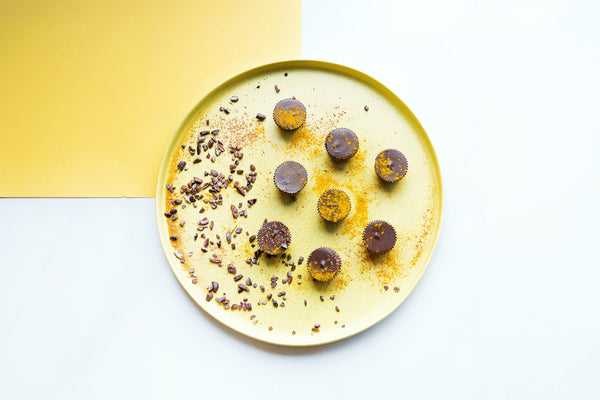 Yellow plate sits on a marble counter. There are chocolate cups sprinkled with turmeric powder on the plate and cocoa nibs surrounding that.