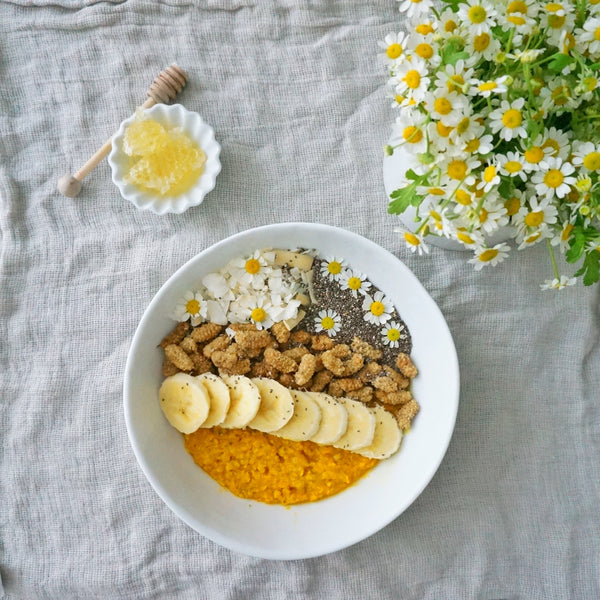 There is a bowl sitting on a white table cloth. There is a bushel of small white and yellow flowers on the table. Inside the bowl there is golden oatmeal, banana slices, nuts, coconut shreds, chia seeds and more flowers like the ones on the table. 