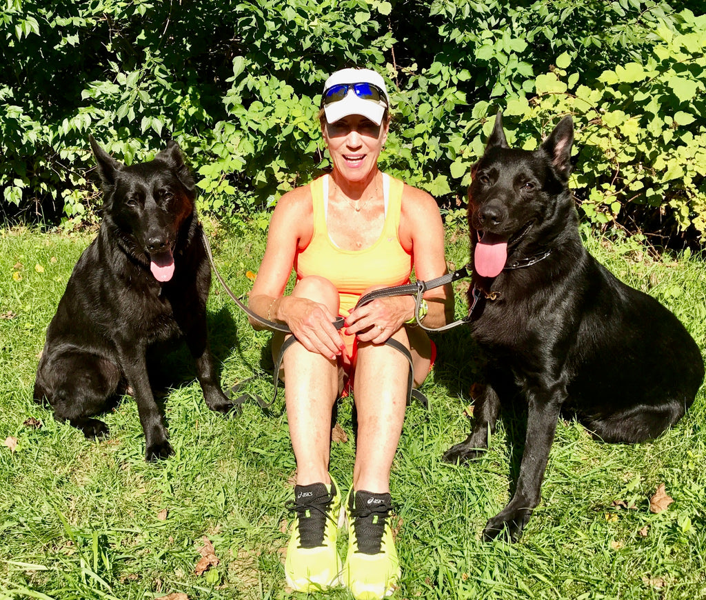 Dr. Pam Peeke in exercise clothes sitting between two large black dogs on a sunny lawn.