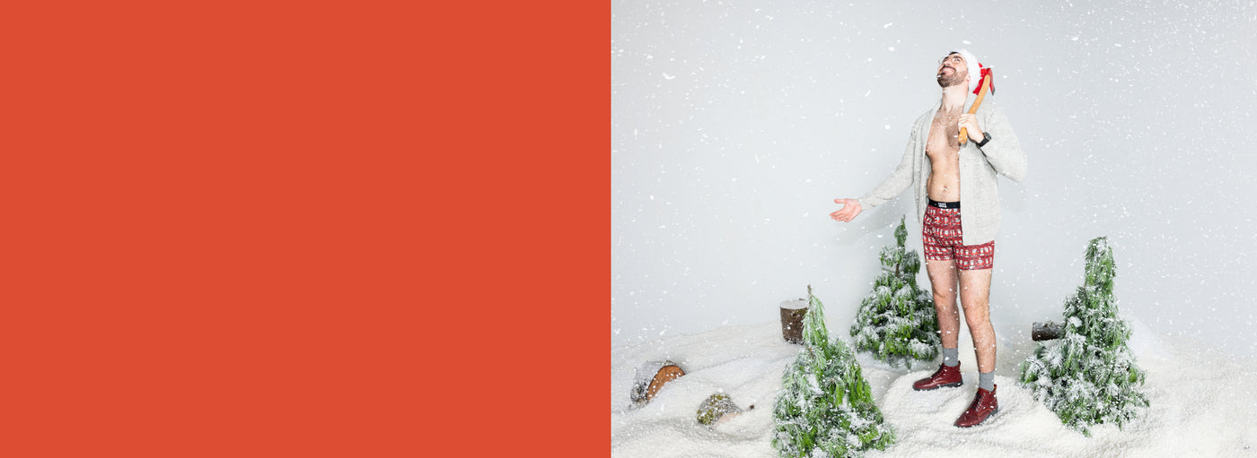 Snow and pine trees surrounding a man holding an ax and wearing Santa hat, red boxer briefs in Santa print, and red boots