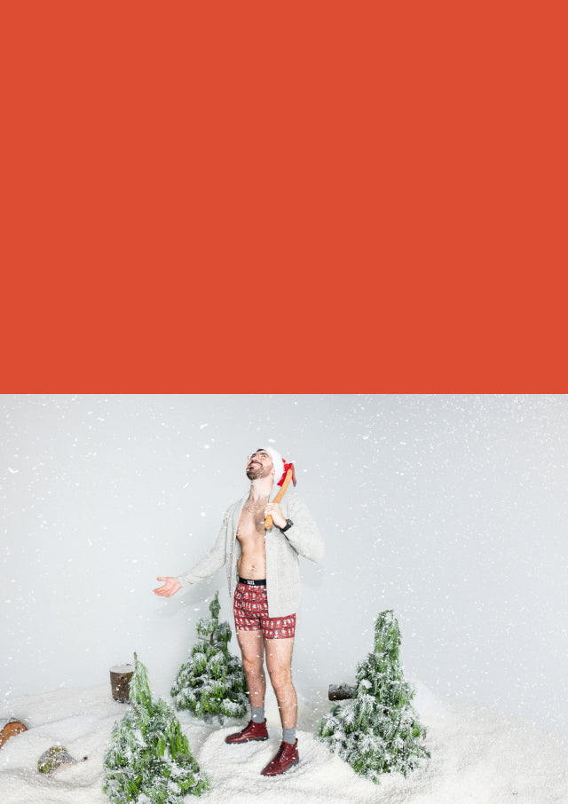 Snow and pine trees surrounding a man holding an ax and wearing Santa hat, red boxer briefs in Santa print, and red boots