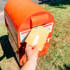 Image of a hand holding a yellow envelope, about to insert the envelope into a bright red mail box by the side of the road. 