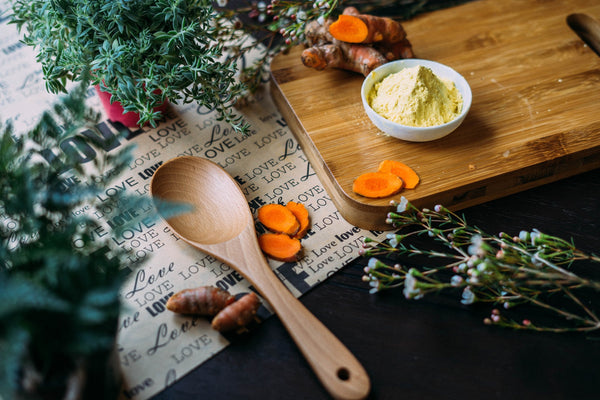 How Ayurveda is useful in modern life. An image of a wooden spoon on a bench beside a some sliced turmeric root and a small bowl of yellow powder and some fresh stalks of green herbs. 