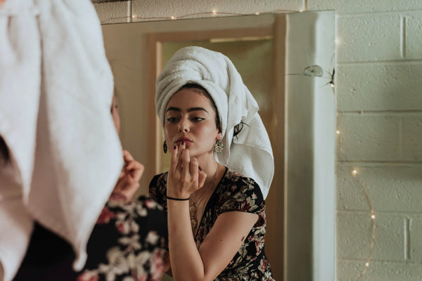 Ayurveda for skincare. A young woman with olive skin and dark features stands in front of a mirror wearing a white towel on her head, as if she's just come out of the shower, and applies balm to her lips. She is wearing a short-sleeved, dark-coloured t-shirt. 