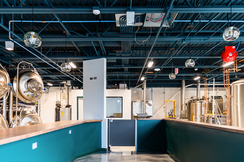 Bicycle Glass Pendant Lighting Spun 768 in clear with single pendant hardware kits in black, featured in a commercial tap room with large brewing machinery, a tap and a bar, deep that shows machinery on the left and right sides of both counters and an open walkway in the middle