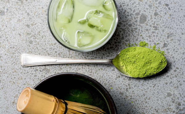 A glass of iced, cold brew matcha tea, a spoon of matcha powder, and traditional preparation bowl.