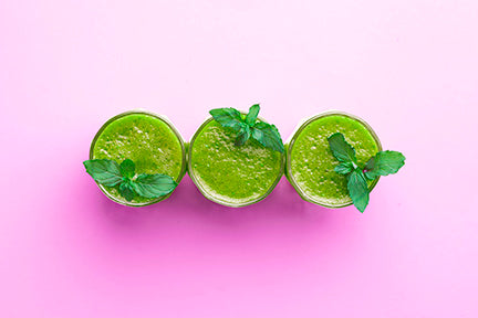 Three glasses of matcha green juice adorned with mint leaves, on a pink background.