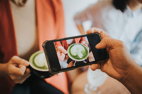 Person taking a photo of a woman's hands holding a matcha latte on a cell phone