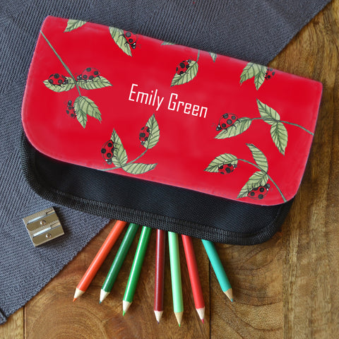 A red personalised ladybird pencil case. The pencil case is personalised with the name "Emily Green" and is on a table next to some coloured pencils.