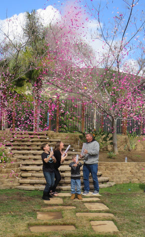 Family posing together outside covered in pink confetti to announce the parents having a baby girl.