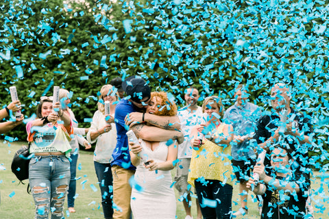 Couple surrounding by family outside, posing with blue confetti to announce having a baby boy