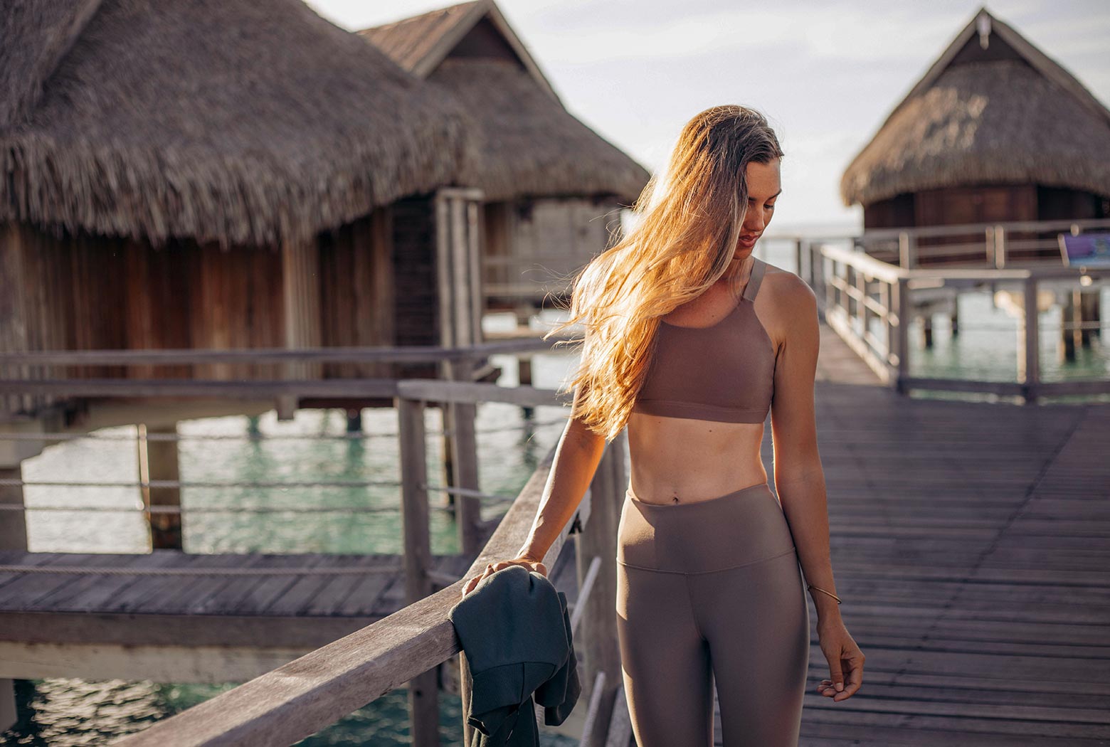 Rachel Moore standing on a polynesian dock in front of tiki huts, wearing a beige sports bra and leggings combo