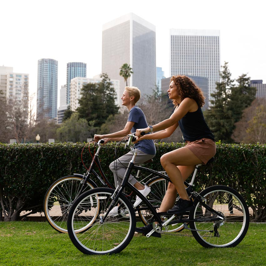 Two Mondetta female models on bicycles, riding through a city park, wearing Mondetta brand activewear
