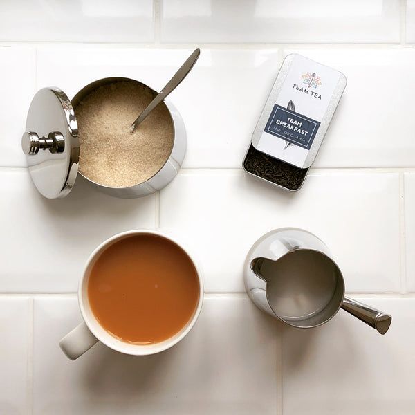 A mug of English breakfast tea, brewed with loose leaves. Along with the mug is a tin of loose leaf tea, a pot of sugar and a jug of milk. The background is white tiles.