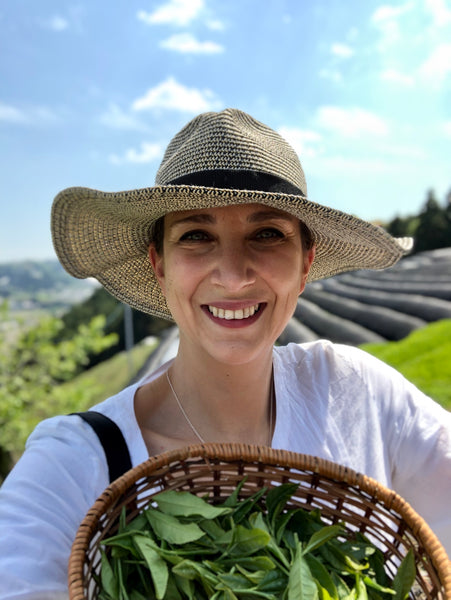 A photo of our founder, Valerie, picking tea leaves in Japan. Valerie is wearing a hat, with beautiful blue sky overhead and a basket of tea leaves in hand.