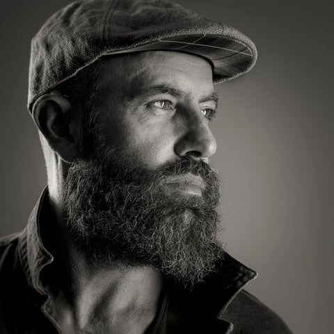 A black and white shot of a man with a thick beard gazing to the right, wearing a cap.