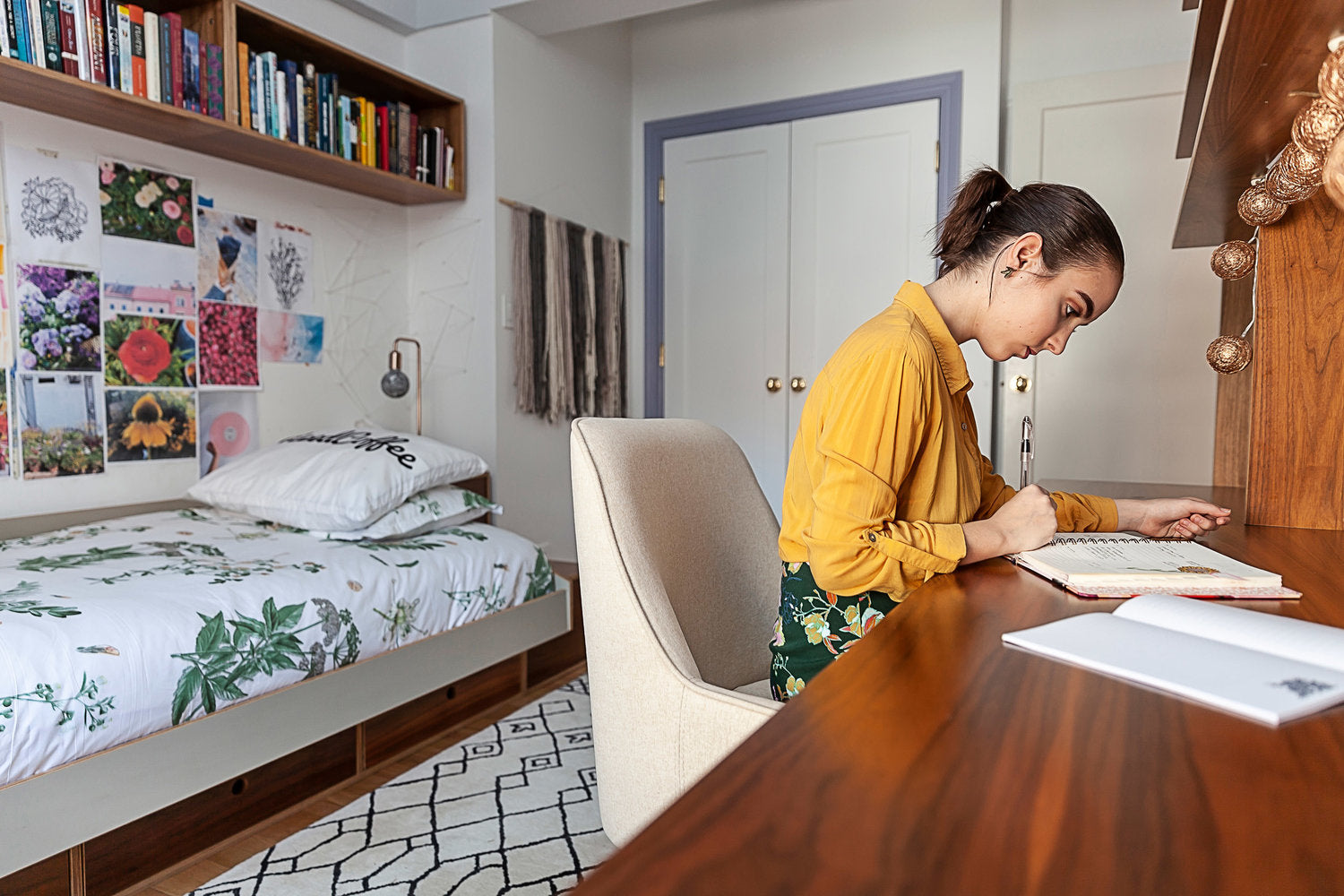 Young woman in yellow shirt studying at a desk in a cozy bedroom with bookshelves and art on walls.