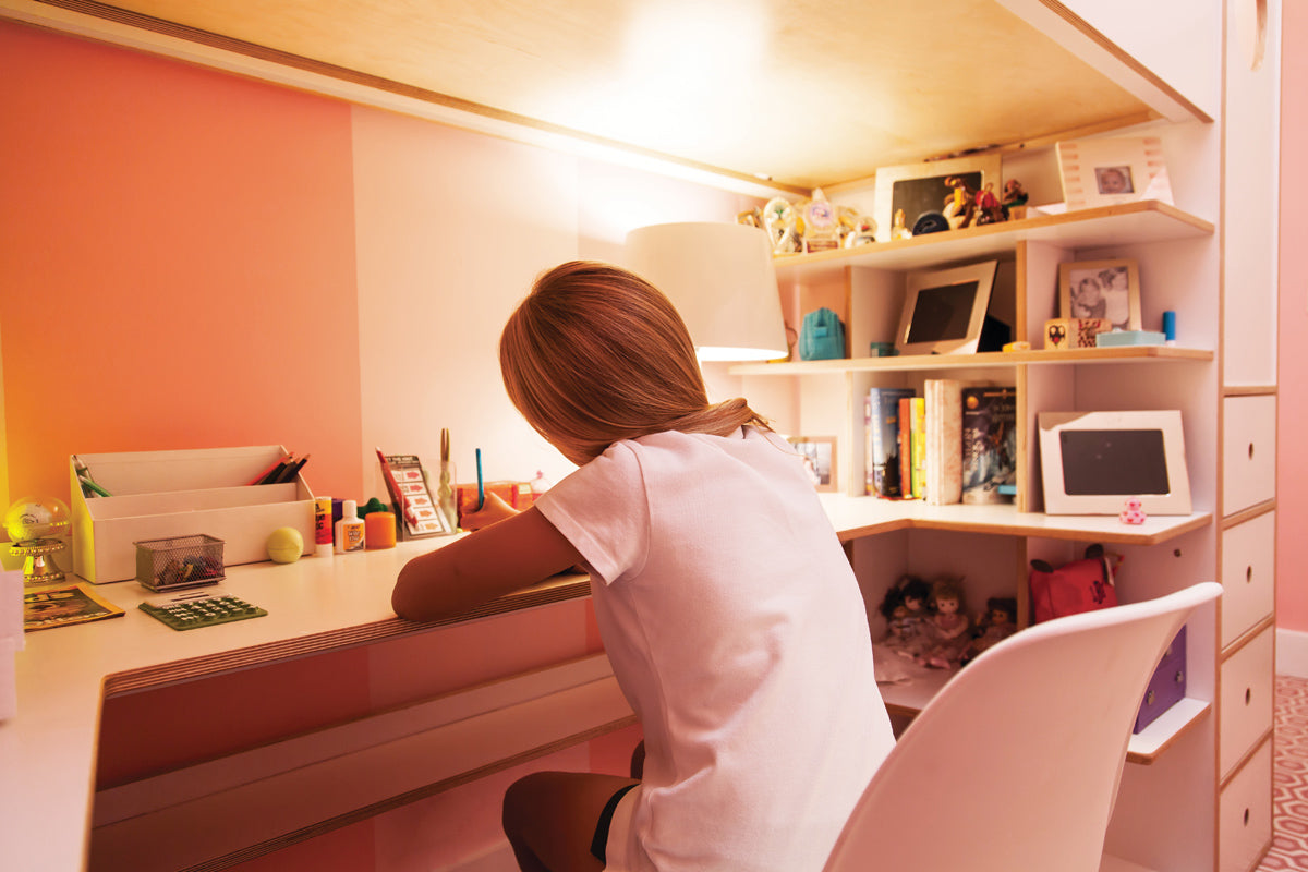Girl studying at a pink desk under a slanted ceiling, surrounded by shelves and warm lighting.