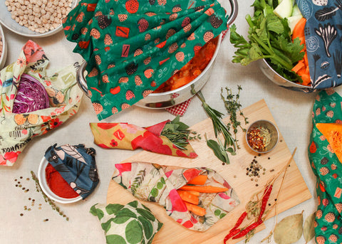 A flatlay image of the construction of a pot of shakshuka, with a variety of ingredients both wrapped and unwrapped scattered across a beige table cloth. Left to right starting at the top: A pale blue bowl of chickpeas; a pot of shakshuka mostly covered by a Strawberry Fields print wrap; a stainless steel bowl of prepped veggies sticking out from under a blue Petals and Pods print Z Wrap; next row from left: a portion of purple cabbage in a Bees Love These print wrap; a small bundle of fresh herbs in a small Painted Poppy wrap; loose herbs and spices on and around a cutting board; bottom most row from left: some loose peppercorns and a sprig of thyme next to a white ramekin of tomato paste partly covered by a blue Petals and Pods wrap; something unseen covered completely by a Leafy Green print wrap; a bunch of carrot sticks in a cone of Farmer's Market print wrap; some dried chili peppers on their branch; a loose bay leaf; half a butternut squash, insides visible, in a Strawberry Fields wrap.