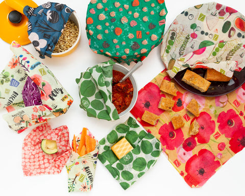 A flatlay image on a white background of colorful Z Wraps containing a variety of items. Clockwise from top right are: a yellow enamel saucepan of pine nuts covered by a blue Petals and Pods print Z Wrap; a large pot totally covered by a green and pink Strawberry Fields print; a dark purple dinner plate with chunks of bread peeking out, covered by a Farmers Market print Z wrap; a flat Painted Poppies wrap with crouton-sized pieces of bread on it; a piece of smoked cheese in a Leafy Green print Z Wrap; carrot sticks in a cone of Bees Love These print wrap; half an avocado in a red and white Connect the Dots print wrap; half a purple cabbage wrapped in Bees Love These floral print wrap; and a bowl of tomato-based food with a spoon in it, half covered by a Leafy Green print wrap in the center.
