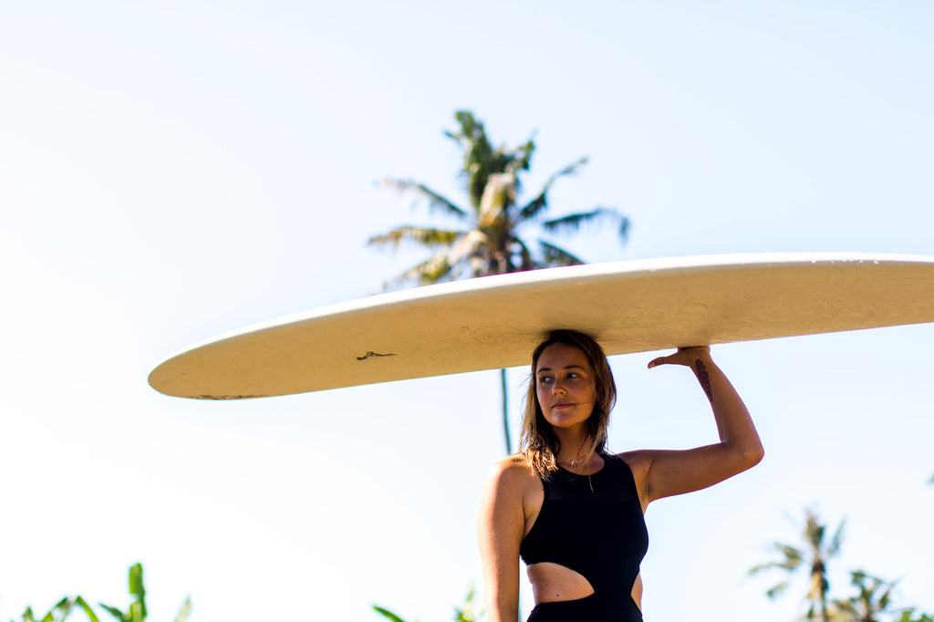 Sierra walking through the rice fields, carrying her surfboard on her head, wearing the Malibu one piece surf swimsuit in black 