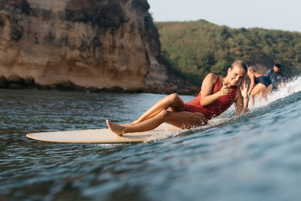 Woman laying down on a surfboard and smiling wearing a one piece surf swimsuit in burnt sienna