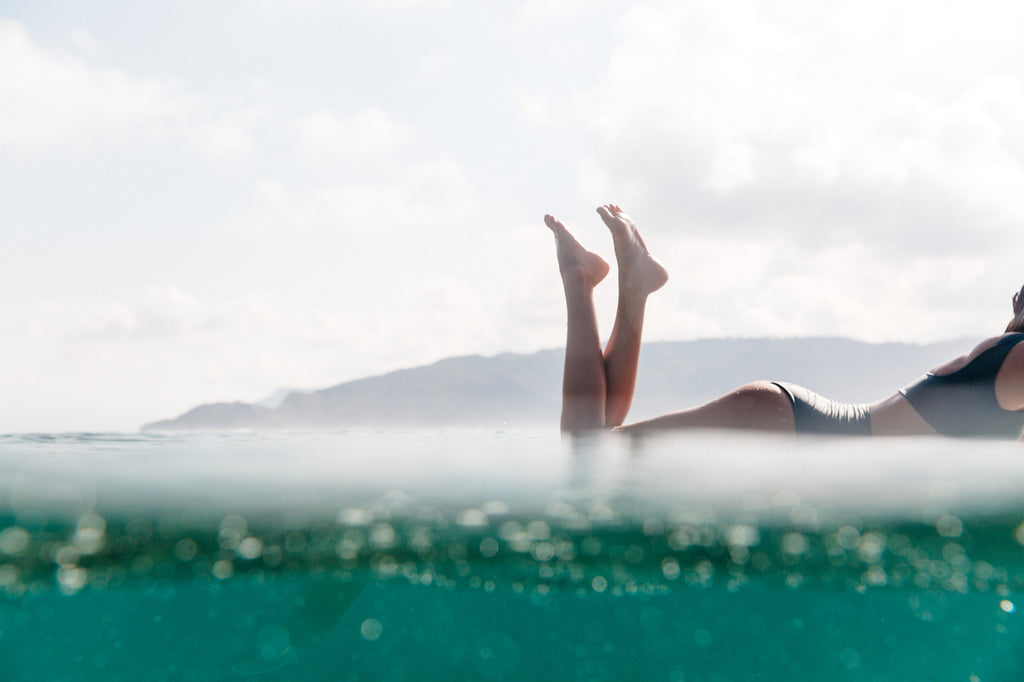 Water photo of woman laying on a surfboard with her legs bent wearing a high waisted surf bikini bottom