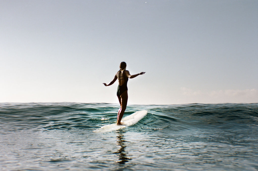 Sierra standing on her board going over a wave wearing a one piece surf swimsuit