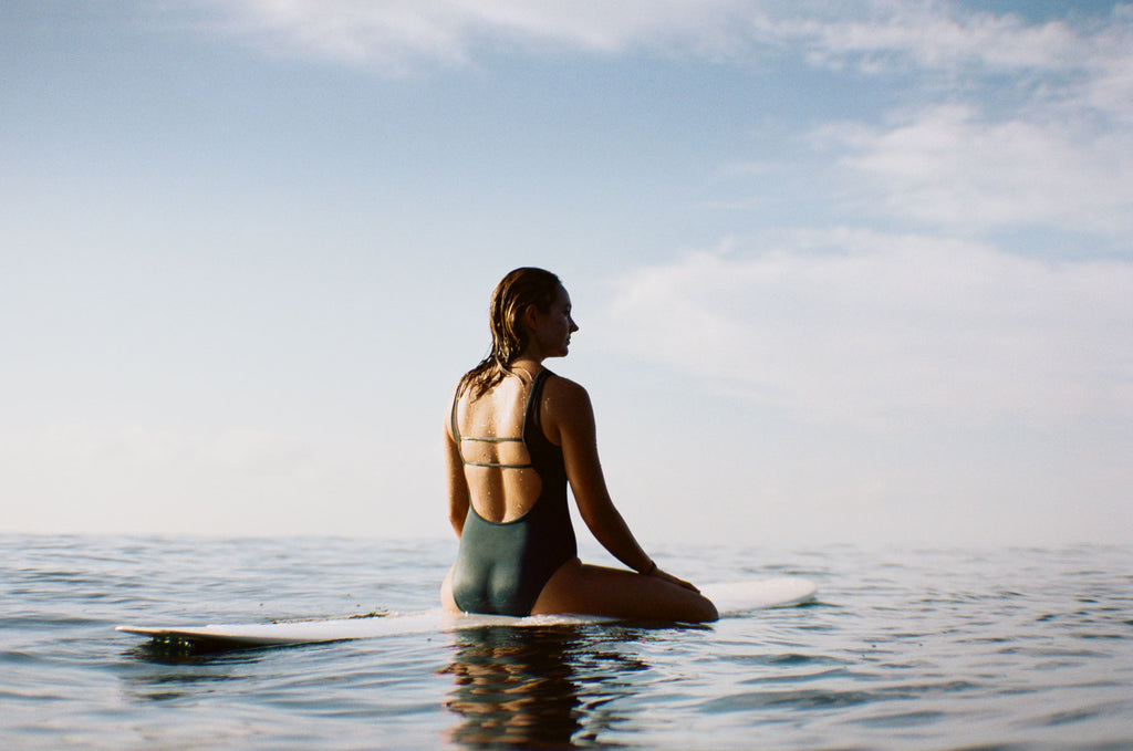 Sierra sitting on her board looking towards the horizon, wearing a one piece surf swimsuit