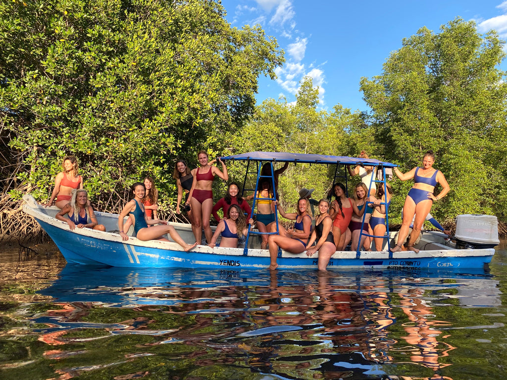 Group of 15 women wearing surf bikinis and one piece swimsuits standing and sitting on fishing boat in the mangroves