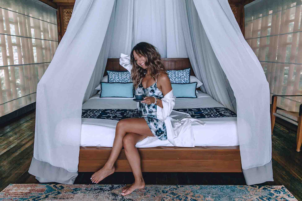 This Island Life wearing the London Print Slip and Ivory Silk Cotton Robe seated on a bed in a beach bungalow on Bawah Island